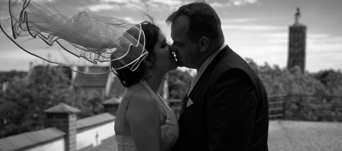 schwarz weiß Fotografie eines Hochzeitspaares auf einer Anhöhe mit wehendem Schleier im Wind und einer Kirche im Hintergrund