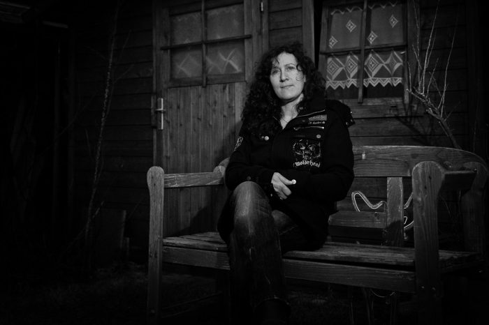 eine Frau sitzt vor einer Holzhütte auf einer Band und blickt in die Kamera